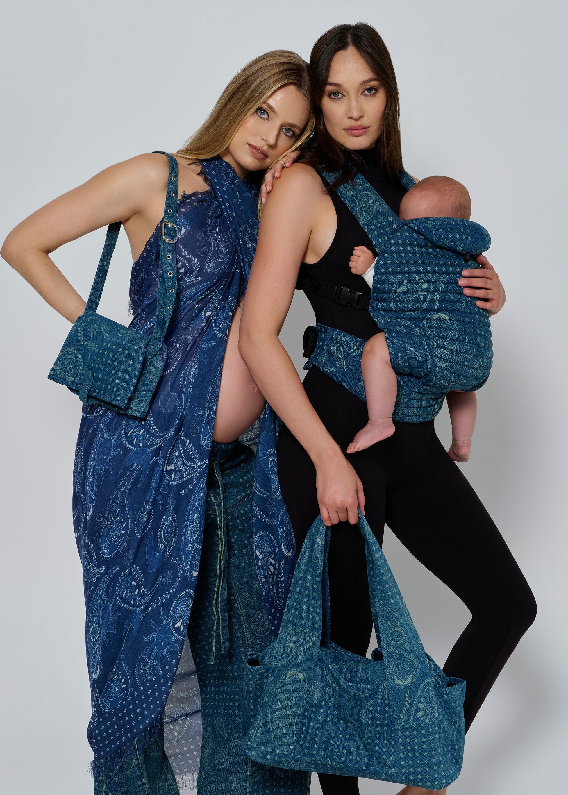 Two women holding blue spell and bumpsuit  textured bags with a baby in a carrier against a plain background