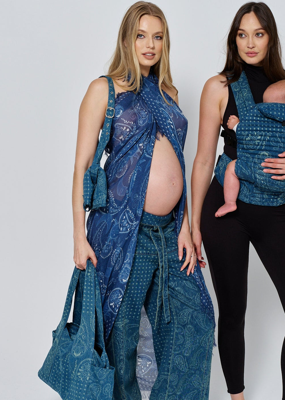 Two women wearing spell print denim outfits with a bumpsuit baby carrier and handbag on a white background