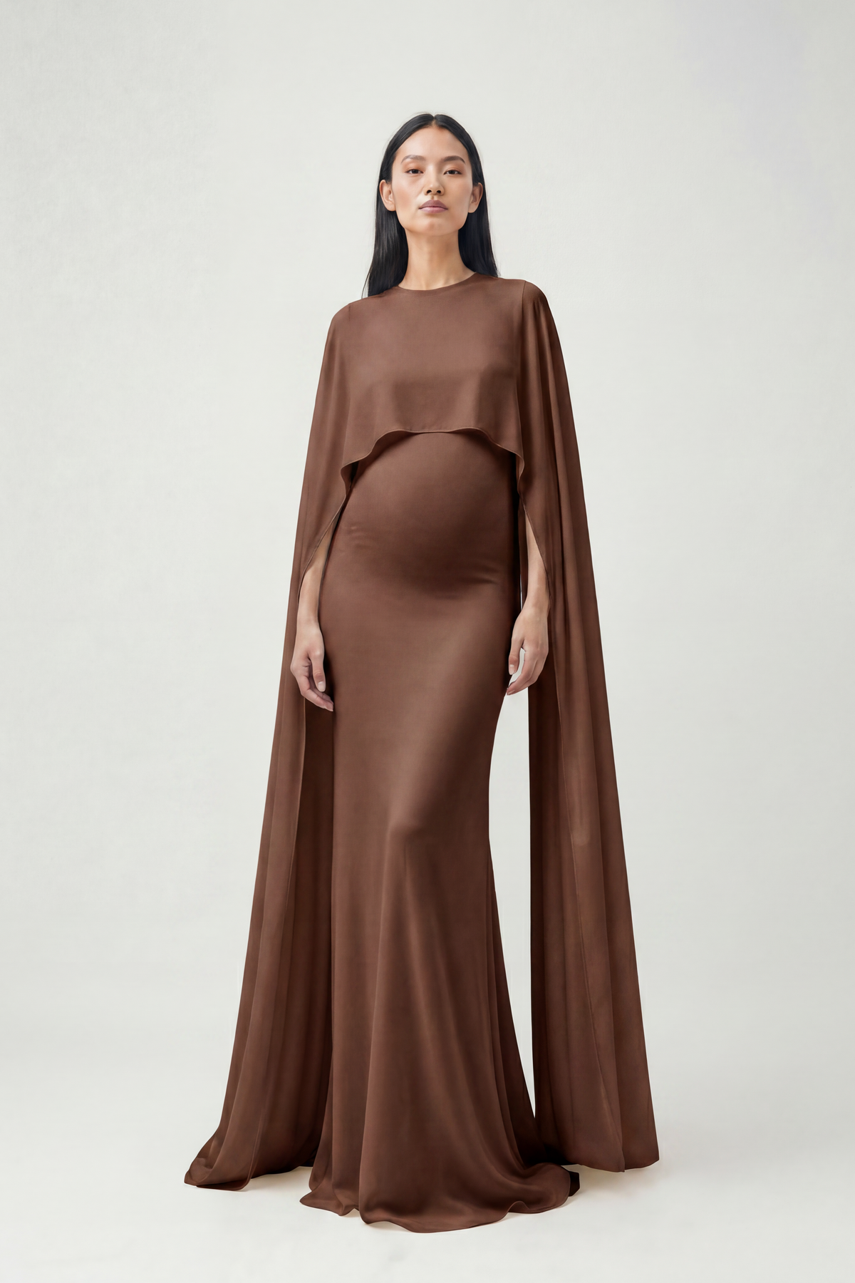 A pregnant model wearing a Bumpsuit brown maternity gown with a draped cape overlay, standing against a light gray studio background.