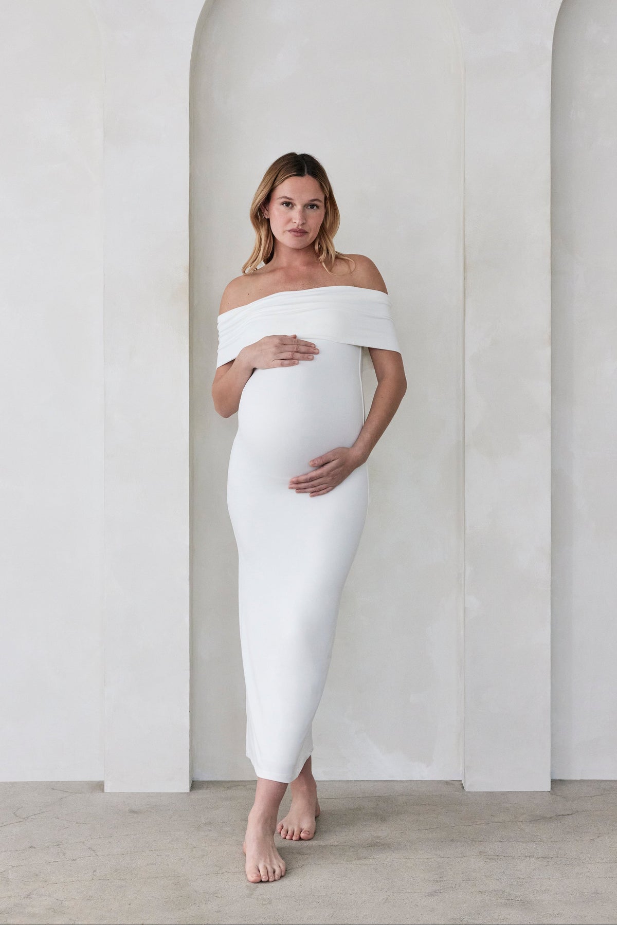 Pregnant woman wearing Bumpsuits off the shoulder Ivory Bianca maternity maxi dress standing  against a beige wall