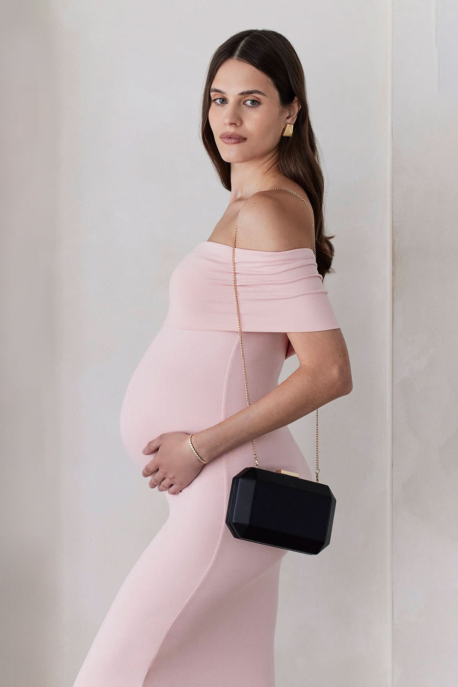 Pregnant woman wearing the Bumpsuit Maternity The Bianca Off Shoulder Maxi Dress in Dusty Pink with a little black bag