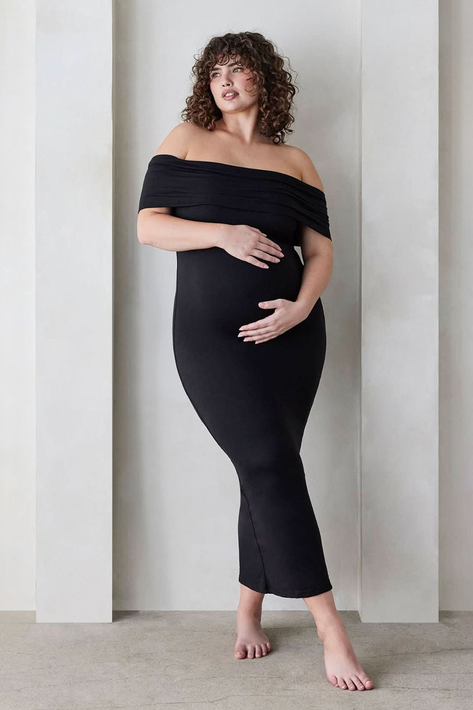 Pregnant woman wearing a Bumpsuit Maternity The Bianca Off Shoulder Maxi Dress in Black standing against a beige wall