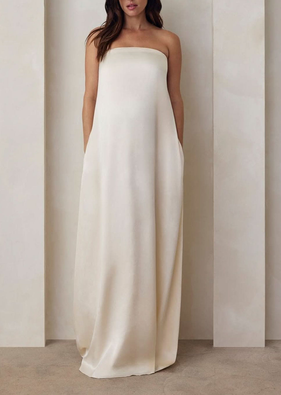 Pregnant model wearing the Bumpsuit brushed satin gown in ivory against a beige feature wall.