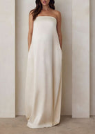 Pregnant model wearing the Bumpsuit brushed satin gown in ivory against a beige feature wall.