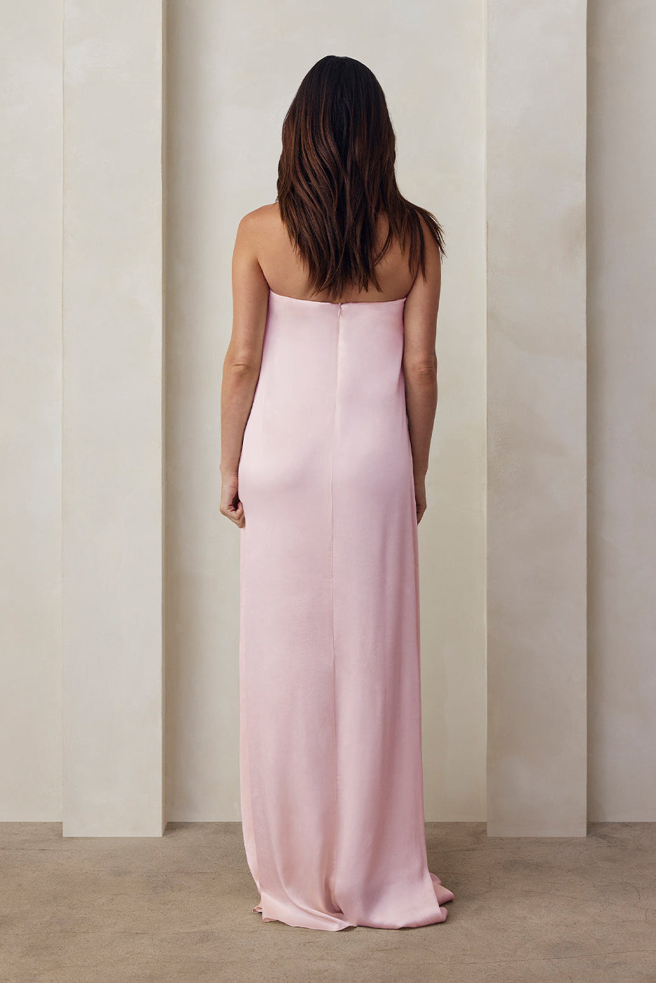 the back view of a pregnant woman wearing the Bumpsuit brushed satin strapless maxi maternity gown in dusty pink against a beige feature wall