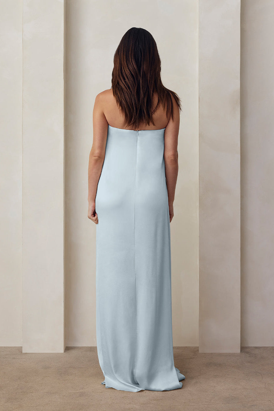 the back view of a pregnant woman wearing the Bumpsuit brushed satin strapless maxi maternity gown in powder blue against a beige feature wall