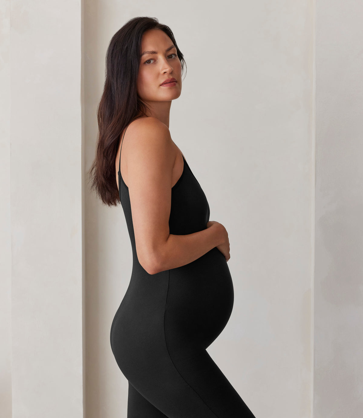 Pregnant woman wearing the Bumpsuit Kate Maternity Jumpsuit in Black against against a beige feature wall