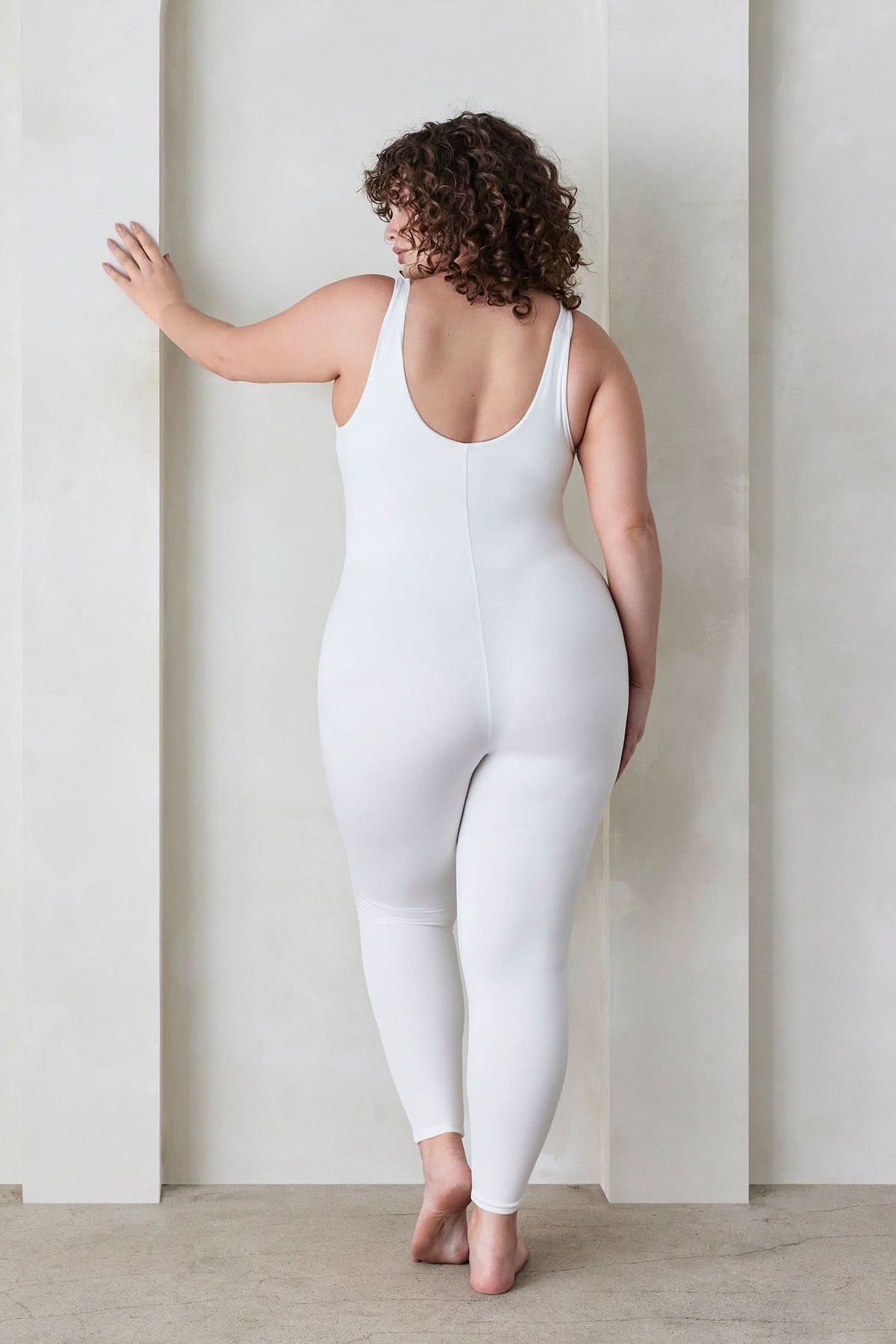 Back view of a curvy woman wearing the Lucy maternity scoop neck unitard in Ivory by Bumpsuit set against a feature wall