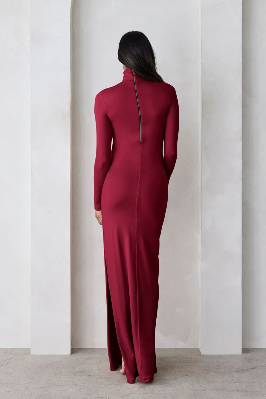 Back view of a pregnant woman wearing a burgundy turtleneck maternity maxi dress with long sleeves and a thigh-high slit on one side standing against a beige feature wall
