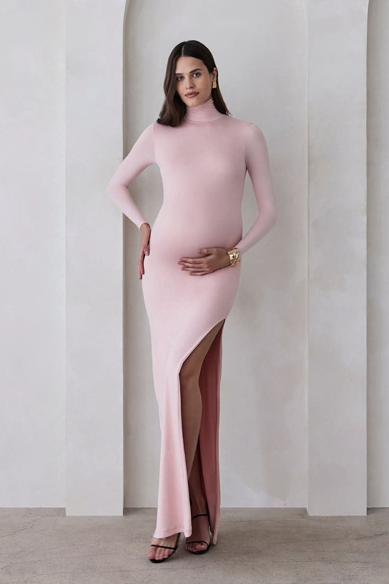 A pregnant woman wearing a dusty pink turtleneck maternity maxi dress with long sleeves and a thigh-high slit on one side standing against a beige feature wall