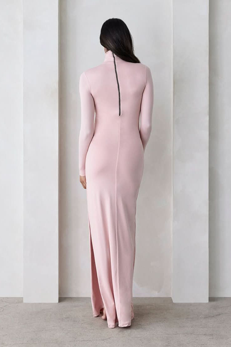 Back view of a pregnant woman wearing a dusty pink turtleneck maternity maxi dress with long sleeves and a thigh-high slit on one side standing against a beige feature wall