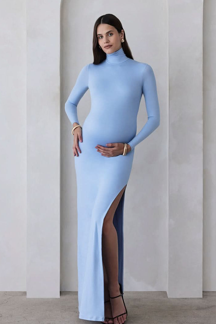 A pregnant woman wearing a powder blue turtleneck maternity maxi dress with long sleeves and a thigh-high slit on one side standing against a beige feature wall