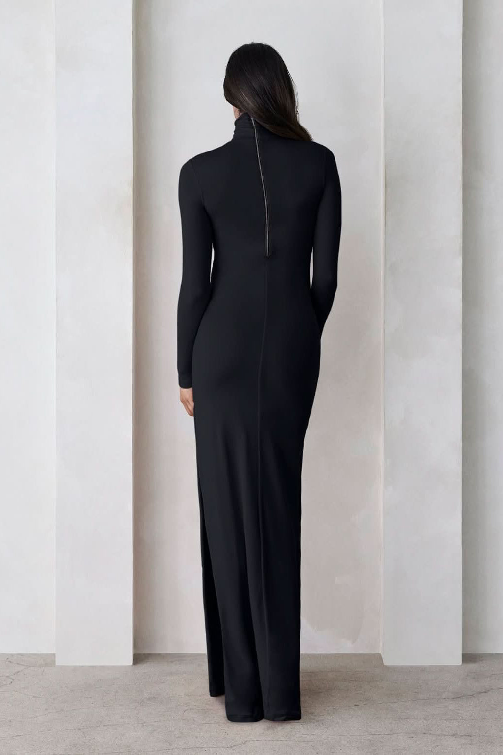 A pregnant woman wearing a black turtleneck maternity maxi dress with long sleeves and a thigh-high slit on one side standing against a feature wall
