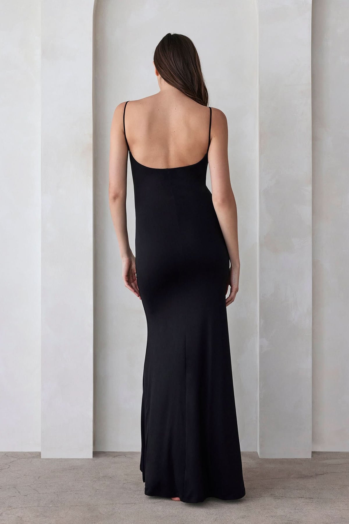 back view of a pregnant woman wearing The Rosie maternity Square Neck Sleeveless Maxi Dress with side slit in black by Bumpsuit,  standing beige against a feature wall
